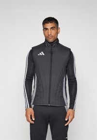 Chaleco atlético negro con cuello alto, cierre frontal, diseño acolchado, paneles laterales grises y acentos en blanco con tres rayas en las mangas.