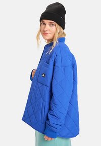 Prošívaná modrá bunda s diamantovým vzorem, s náprsní kapsou a žebrovanými manžetami. Nošena s černou čepicí.