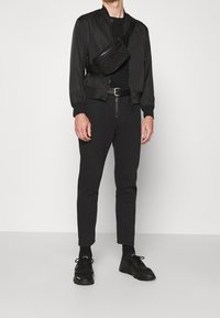 Mężczyzna w czarnej kurtce bomber, czarnym t-shircie, slim fit czarnych spodniach, czarnym pasku, czarnych sneakersach i pikowanej czarnej torbie na ramię na białym tle.