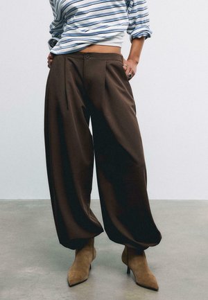 Personne portant un pantalon ample marron foncé, des bottes à talons pointues couleur fauve et une chemise rayée à manches longues avec manches retroussées.
