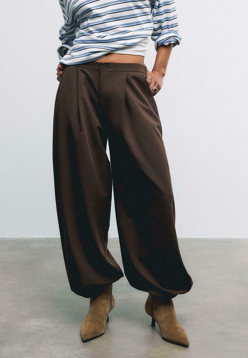 Personne portant un pantalon ample marron foncé, des bottes à talons pointues couleur fauve et une chemise rayée à manches longues avec manches retroussées.