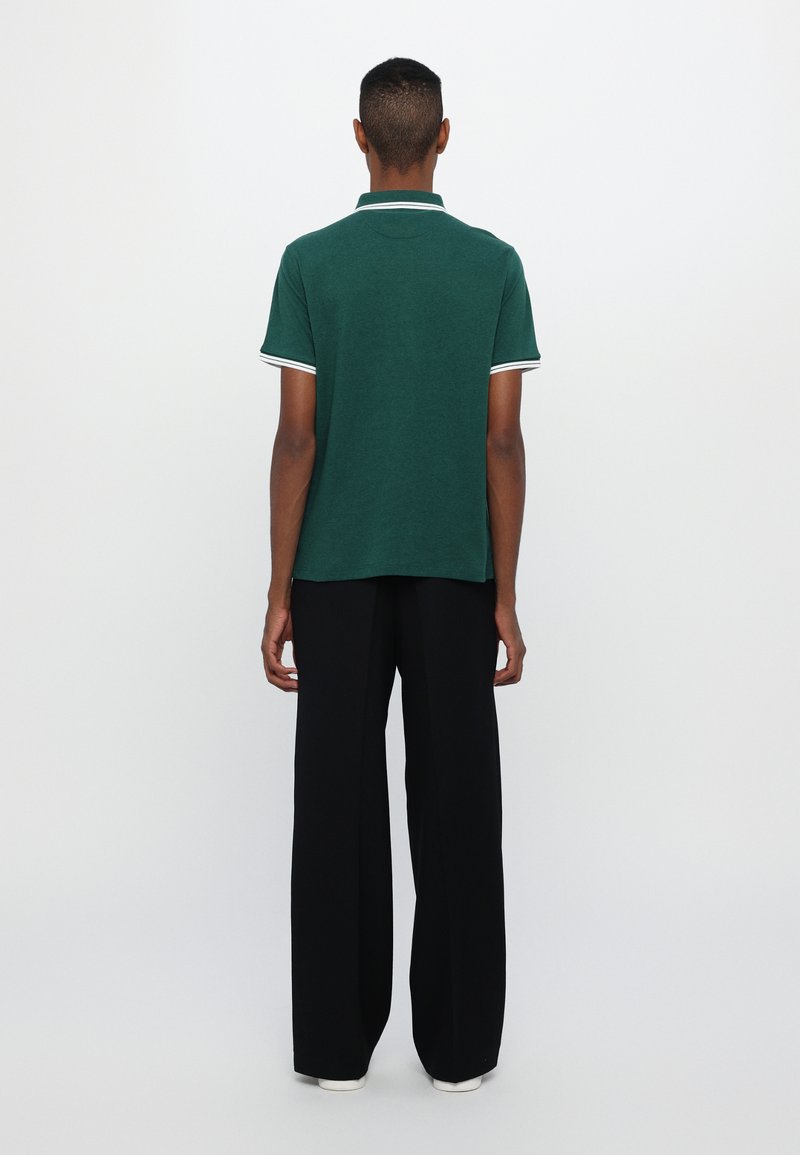 Grünes Poloshirt mit weißem Kragen und Ärmelbesatz, kombiniert mit schwarzen weit geschnittenen Hosen. Glattes Material, klassisches Design, Rückansicht.