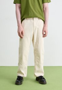 Pantalon en coton beige avec poches latérales, coupe décontractée et fermeture à bouton, associé à des chaussures noires avec une semelle texturée et des franges.