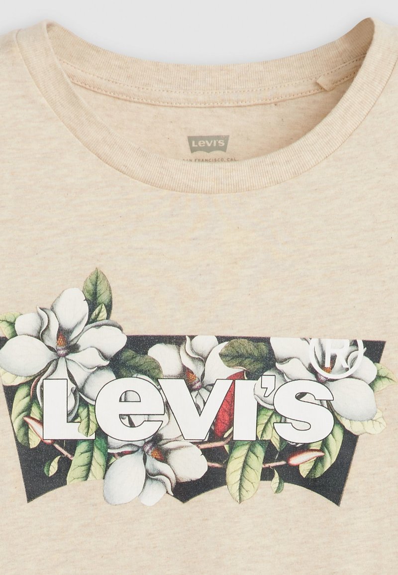 T-shirt en coton beige avec une illustration florale comprenant des fleurs de magnolia blanches, des feuilles vertes et un logo central "Levi's" en blanc.
