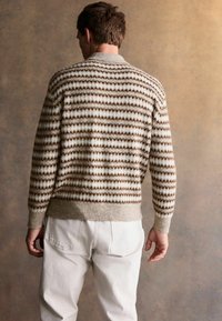 Beige und cremegfarbener gestrickter Pullover mit braunem Zickzackdesign; verfügt über einen gerippten Kragen und Bündchen. Getragen über weißen Hosen.