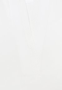 Biała bawełniana bluzka z dekoltem w kształcie V, wykończona prążkowanym brzegiem. Gładka faktura, czysty, minimalistyczny wygląd bez widocznych wzorów.