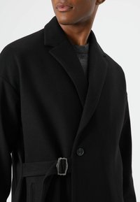 Schwarzer Wollmantel mit Reverskragen, Knopfverschluss und einem silbernen Schnallendetail an der Taille. Glatte Textur mit tailliertem Schnitt.