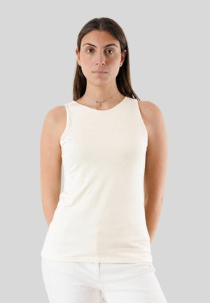 Donna con lunghi capelli scuri che indossa una canottiera bianca senza maniche e pantaloni bianchi, in piedi davanti a uno sfondo grigio uniforme.