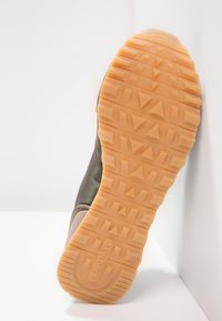 Schoen met een textuur in olijfgroen bovenwerk, rubberen zool in tan met geprononceerde grippatronen en logo aan de zijkant.