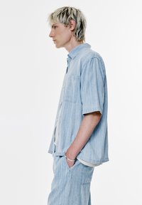 Camicia azzurro chiaro a maniche corte con righe verticali, apertura con bottoni, taschino sul petto, indossata con pantaloni abbinati di un blu chiaro più intenso.