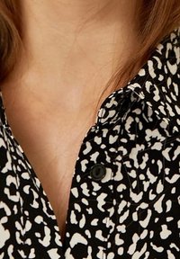Camisa de patrón en blanco y negro con cuello tipo solapa, que presenta formas abstractas y un detalle de botón en el cuello. Textura de tela suave.
