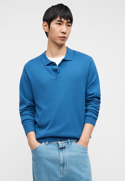 Μπλε πόλο μπλούζα με μακριά μανίκια, γιακά, πλάκα με δύο κουμπιά και ριμπ μανσέτες, συνδυασμένη με ανοιχτόχρωμο μπλε τζιν.