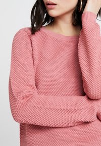 Maglione lavorato a maglia rosa chiaro con un motivo testurizzato, scollatura rotonda e maniche lunghe. Presenta polsini a costine e una vestibilità rilassata.