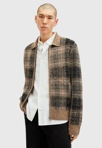 Brun og sort ternet cardigan, lavet af blød uldblanding, med lynlås foran og en beige krave, båret over en hvid skjorte og mørke bukser.