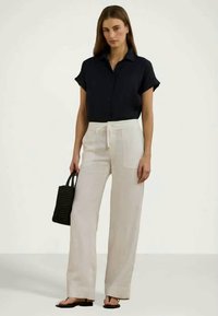 Camisa azul-marinho de manga curta, combinada com calças brancas de perna larga, uma bolsa de mão trançada preta e sandálias pretas, contra um fundo neutro.