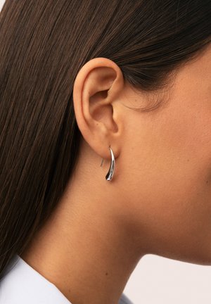 Primer plano de la oreja de una persona con cabello castaño liso, que lleva un pequeño aro plateado brillante.