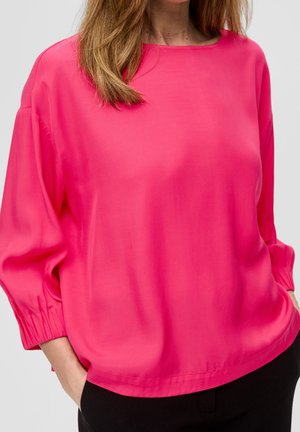 Kobieta ubrana w jaskrawą różową bluzkę z długim rękawem z marszczonymi mankietami i czarne spodnie, ręce w kieszeniach, na jasnym tle.