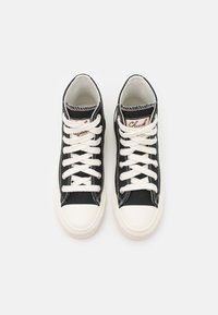 Para czarnych wysokich trampków z płótna z białymi gumowymi noskami, białymi sznurowadłami i różowymi metkami na języku, widziana z góry na białym tle.