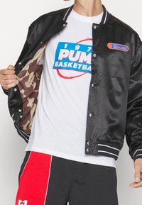 Black satin varsity jacket with white striped collar and cuffs, featuring a camouflage lining. White t-shirt with blue and red "PUMA BASKETBALL" graphic, red shorts.