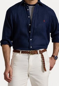 Marineblauw linnen overhemd met witte knopen, gestreept ondershirt, gevlochten bruine leren riem, witte broek en een blauw horloge.