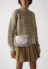 Crossbody torba iz svetlo sive tkanine, z gladko teksturo, detajli z zadrgo in nastavljivim trakom. Nosi se z pjkastim puloverjem.