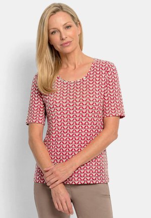 Blonde woman with straight hair wearing a red patterned short-sleeve top and beige pants, standing with hands crossed.