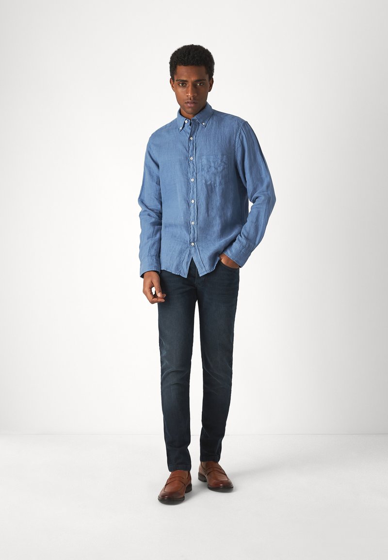 Hellblaues Button-Up-Hemd mit einer Tasche, kombiniert mit dunklen Jeans und braunen Schuhen. Das Hemd hat einen lässigen Schnitt und lange Ärmel.