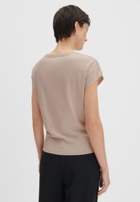 Top beige a maniche corte con colletto rotondo, texture liscia, orlo a costine e una vestibilità comoda. La parte posteriore è leggermente più lunga di quella anteriore.