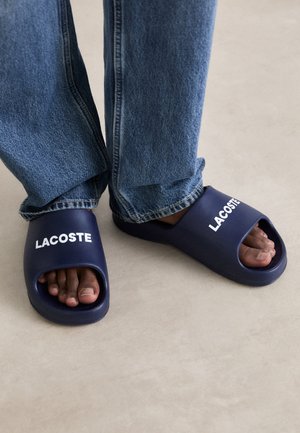 Tamno plave gumene klizne sandale s otvorenim prstima i sa strane, sa bijelim 'LACOSTE' brendiranjem na trakama. Nošene uz traperice.