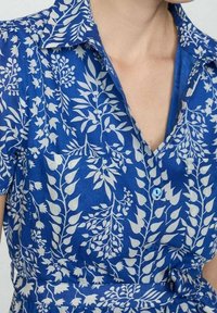 Camicetta blu con motivo floreale bianco, dotata di colletto, maniche corte, chiusura frontale con bottoni e dettaglio in vita annodato.