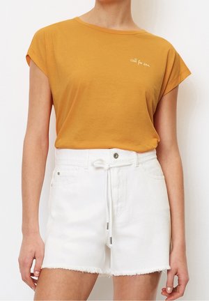 Frau trägt ein kurzärmeliges Oberteil in Senfgelb mit dem Schriftzug "call for sun" und weiße, hoch taillierte Shorts mit Fransen am Saum und Kordelzug.