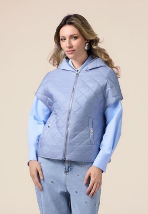 Gepolsterte, hellblaue Kapuzenjacke mit Reißverschluss, weiten Ärmeln und zwei vorderen Taschen. Getragen über einem blauen Shirt und verzierten Jeans.