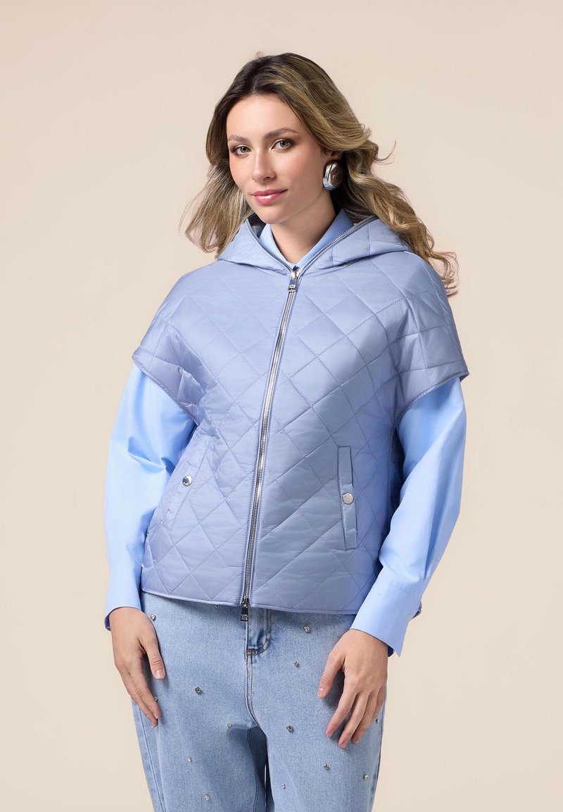Gequilte lichtblauwe hoodie-jack met een rits aan de voorkant, wijde mouwen en twee voorzakken. G gedragen over een blauw shirt en versierde jeans.