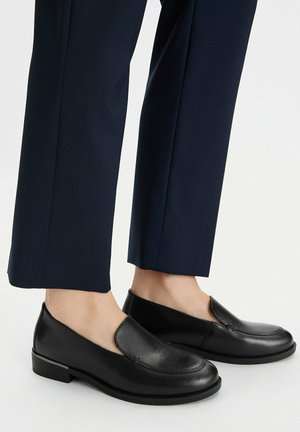 Mocasines negros de cuero sin cordones usados con pantalones a medida de color azul marino, mostrados sobre pies descalzos contra un fondo blanco liso.