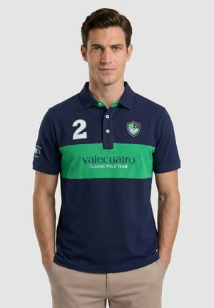 Hombre vestido con camisa polo azul marino con una franja horizontal verde, número 2 en el pecho y pantalones beige, de pie con las manos en los bolsillos.