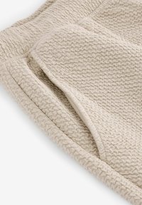 Beige tekstureret stof med et vævet mønster, der har en let plyssurface og en blød, foldet kant. Velegnet til komfortabel brug.