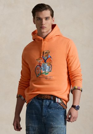 Oranž kapuutsiga dressipluus, millel on kujutatud karu, kes kannab vabaaja riietust ja jalgratast. Kandmiseks koos ruudulise särgi varrukaäärte ja helesiniste teksadega.