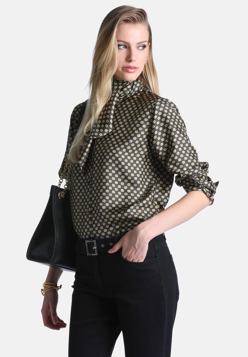 Femme aux longs cheveux blonds portant un chemisier à manches longues à motifs et un pantalon noir, tenant un sac à main noir et regardant sur le côté.
