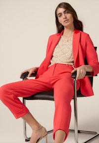 Koraalrode op maat gemaakte blazer en bijpassende cropped broek gecombineerd met een beige pailletten top, gedragen door een zittend persoon op een zwarte stoel.
