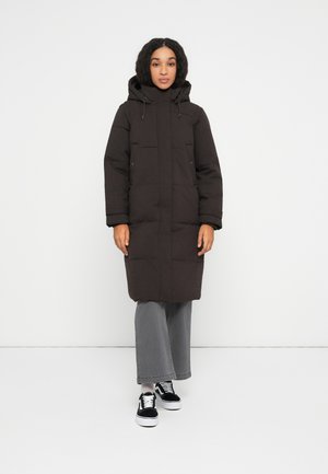 Mujer de pie vestida con un abrigo largo de plumas negro con capucha, pantalones gris de pierna ancha, calcetines blancos y zapatillas de skate negras con cordones blancos.