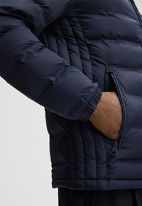 Navy pufferjack met gekwilt ontwerp, textuurstof en zijzakken met rits. Elastische manchetbanden verbeteren de pasvorm en de warmte.