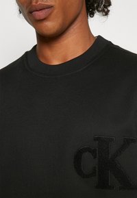 Black cotton sweatshirt featuring a round neckline and ribbed cuffs, with an embroidered "CK" logo in a contrasting texture on the chest.