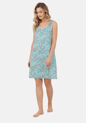 Kobieta stojąca boso, ubrana w turkusową sukienkę w kwiaty bez rękawów, sięgającą kolan, z falbanami przy dekolcie.