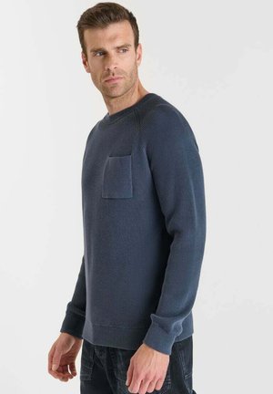 Uomo con maglione blu navy a maglia con tasca sul petto che guarda verso sinistra su sfondo chiaro e uniforme.