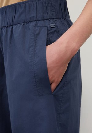 Hånd stukket ind i sidelommen på navyblå elastisk taljebukser på lys-hudet arm og torso.