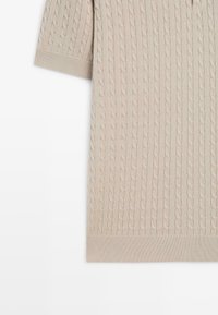 Kabelgebreide korte mouwtrui in lichtbeige met geribbelde zoom- en mouwranden. Heeft een gestructureerd patroon met subtiele contrasterende draden.