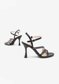 Schwarze High Heels Sandalen mit mehreren gekreuzten Riemen, spitzer Zehenform und Schnallenverschluss. Strukturierte metallische Akzente entlang der Riemen.