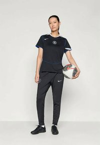 Kobieta stoi, trzymając piłkę nożną, ubrana w czarne ubrania sportowe Nike z niebieskimi akcentami i czarnymi korkami na tle jednolitym.