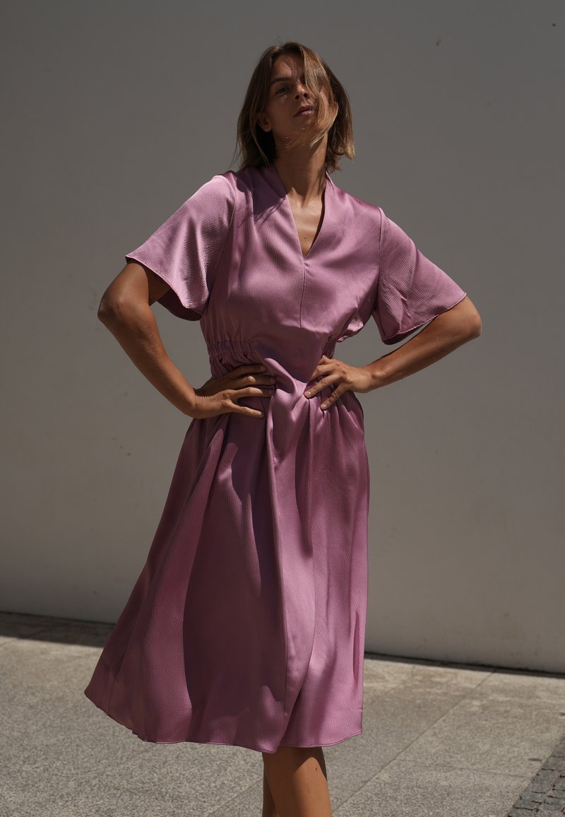 Femme debout, les mains sur les hanches, portant une robe rose ample sur un fond de mur gris uni.