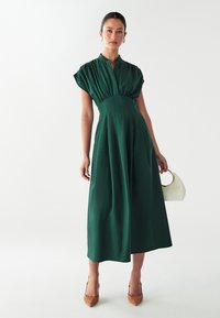 Žalia midi suknelė su trumpomis pūkuotomis rankovėmis, mygtukų apykaklė, prigludusi juosmens dalis ir plačiai krentanti sijonas. Suderinta su balta rankine ir raštuotais bateliais.